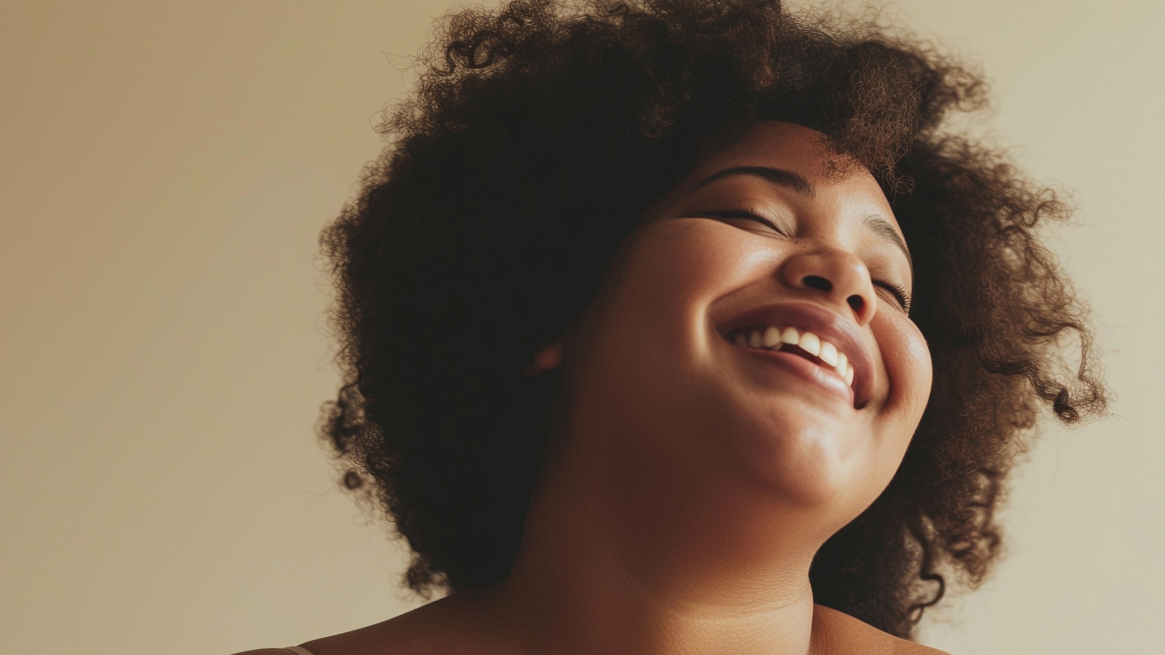 A close-up portrait of a smiling woman with voluminous dark curly hair, wearing a light beige spaghetti-strap top against a warm beige background.