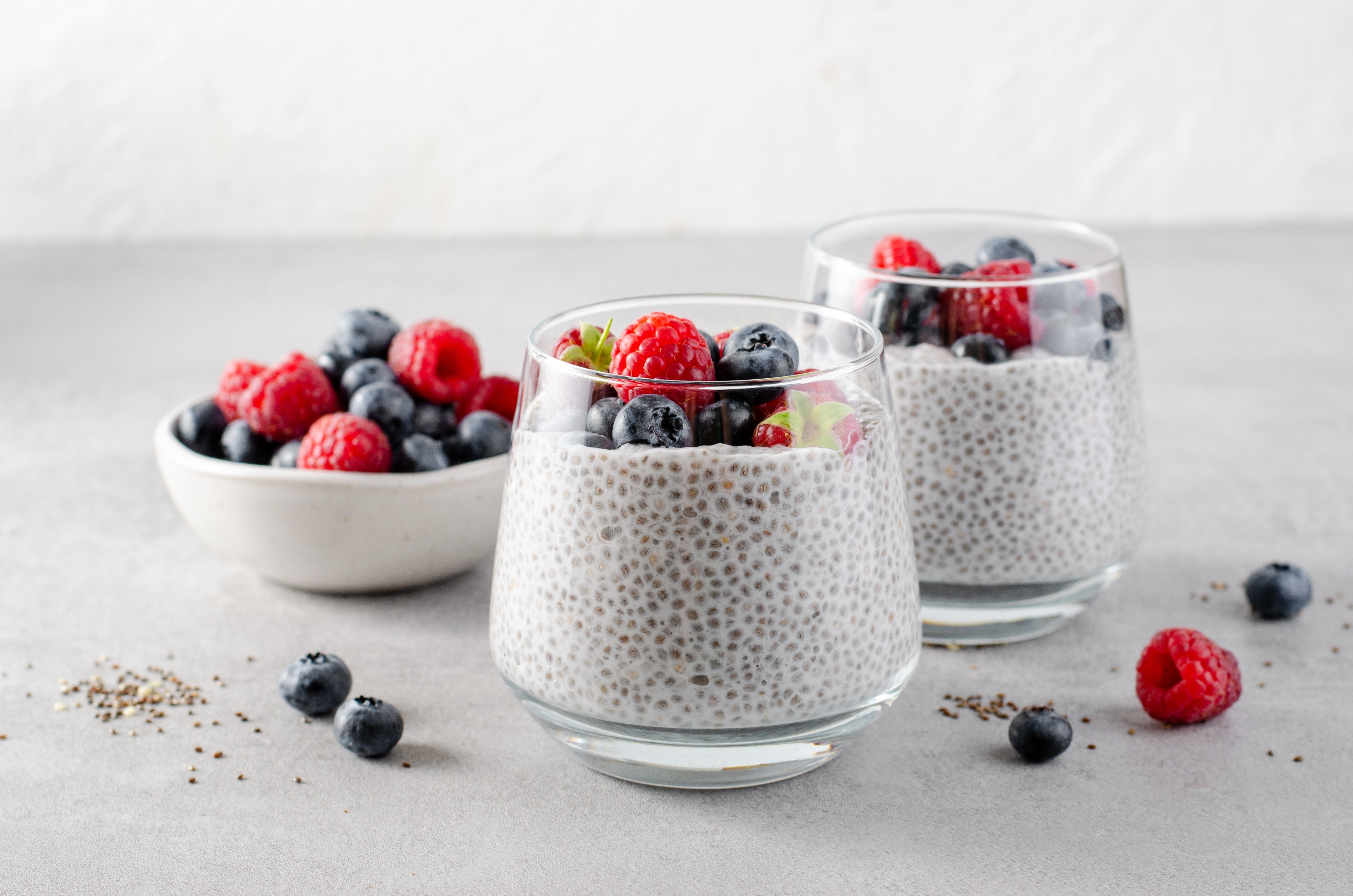 Two clear glass cups filled with chia seed pudding topped with fresh raspberries and blueberries, set on a light gray surface with a small bowl of mixed berries in the background.