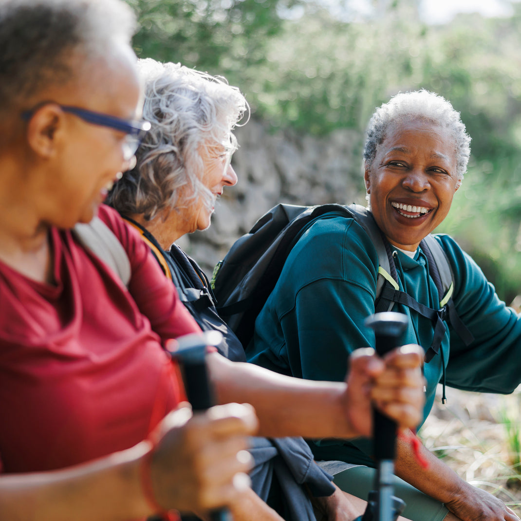 Three smiling senior women hiking outdoors, holding trekking poles. One woman wears a red shirt, another wears a teal shirt with a black backpack, and the third wears glasses and a red top.