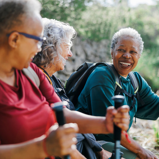 Three smiling senior women hiking outdoors, holding trekking poles. One woman wears a red shirt, another wears a teal shirt with a black backpack, and the third wears glasses and a red top.