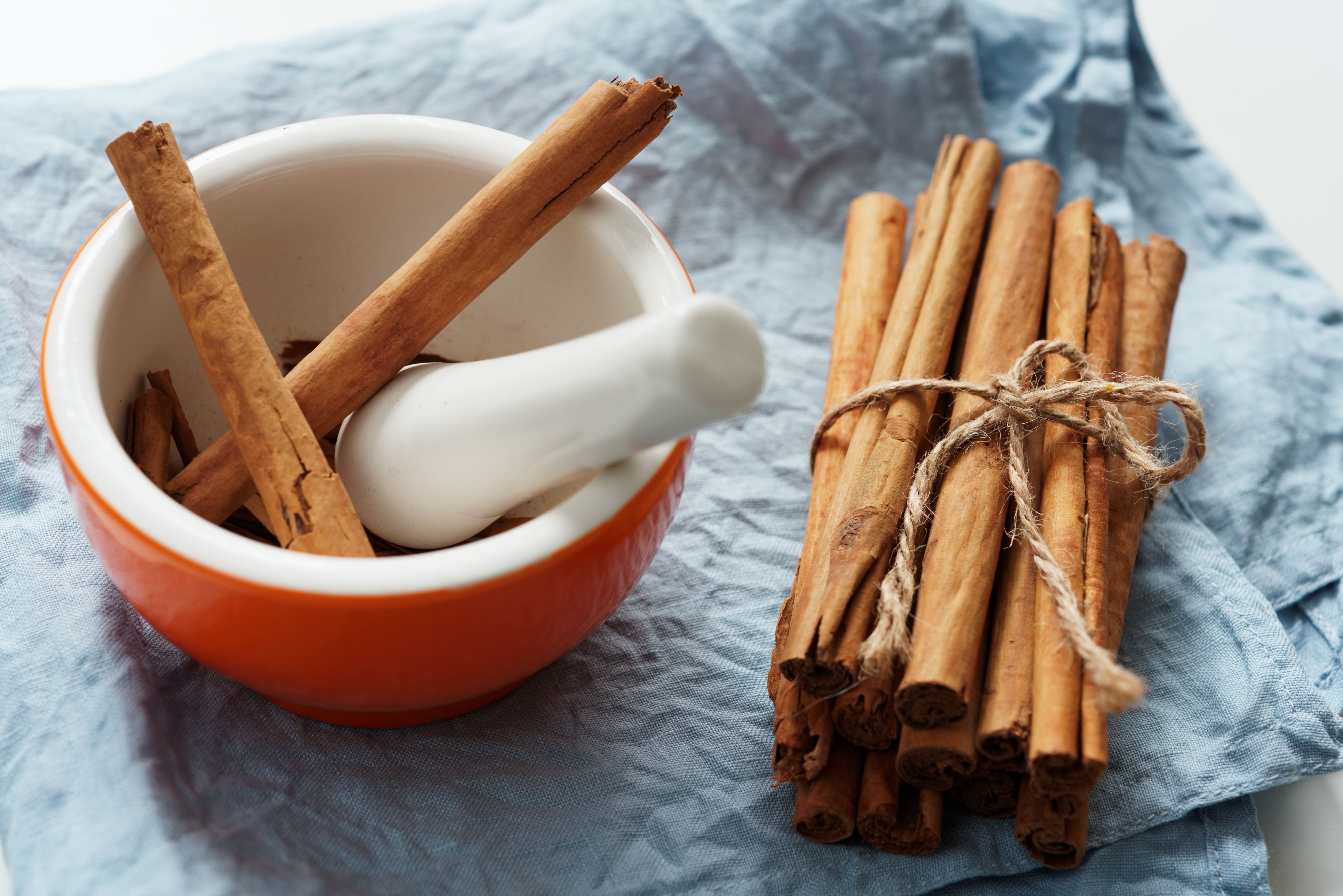 Whole cinnamon sticks, some placed in an orange and white ceramic mortar and pestle and others tied together with twine, set on a crumpled blue cloth.