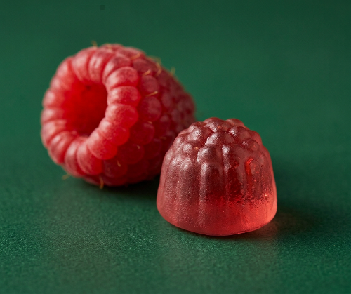 Two red raspberry-shaped gummy candies on a dark green background, with one gummy shaped like a whole raspberry and the other like a halved raspberry.