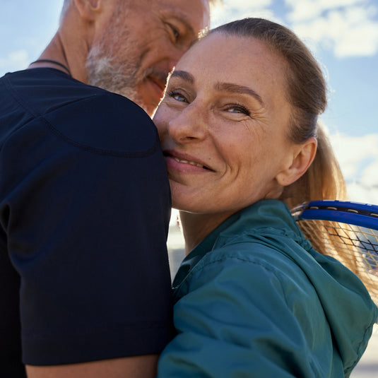 A close-up lifestyle shot of a smiling woman in a teal athletic jacket hugging a man outdoors, with a tennis racket visible behind her shoulder against a bright sky background.