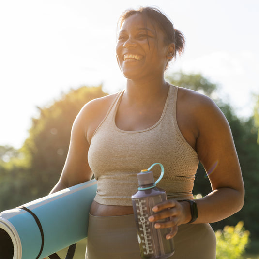A smiling woman outdoors in athletic wear, holding a light blue yoga mat and a gray water bottle. She is wearing a beige sports tank top and matching leggings, with a bright sunlit background.