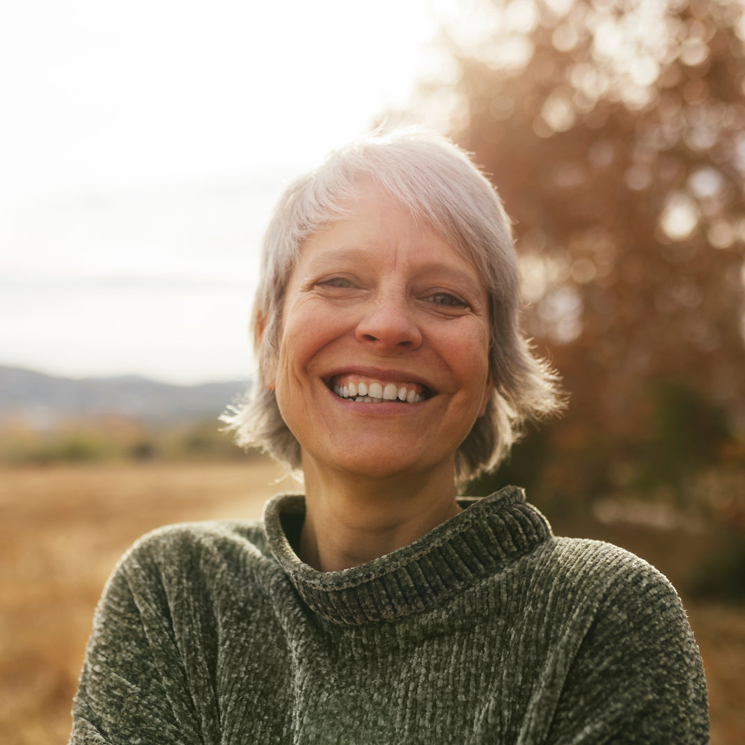 A smiling woman with short gray hair wearing a textured dark green turtleneck sweater, photographed outdoors in warm natural light with a softly blurred field and trees in the background.