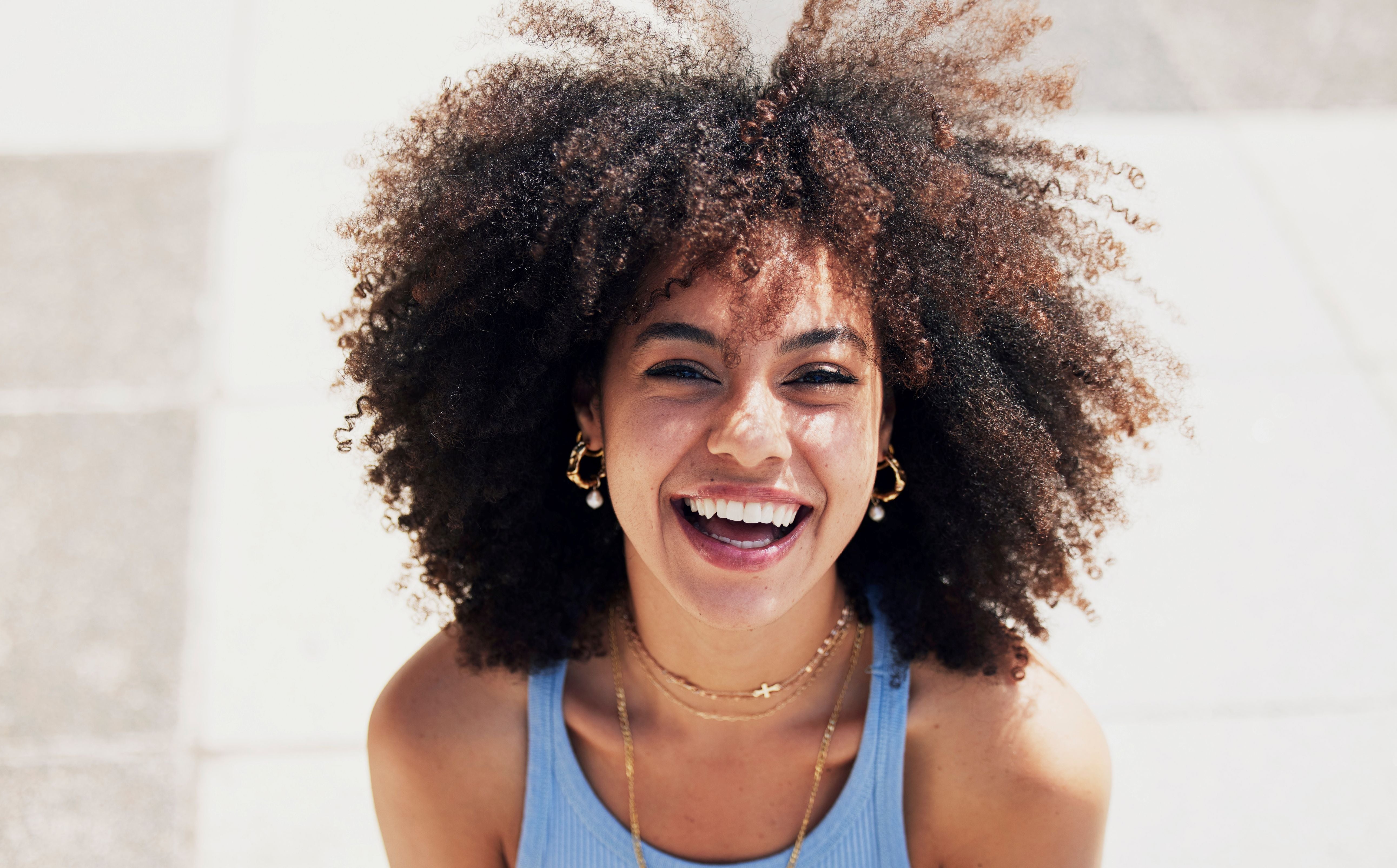 A smiling woman with voluminous dark brown curly hair wearing a light blue tank top and gold jewelry, photographed outdoors against a bright light gray stone wall.