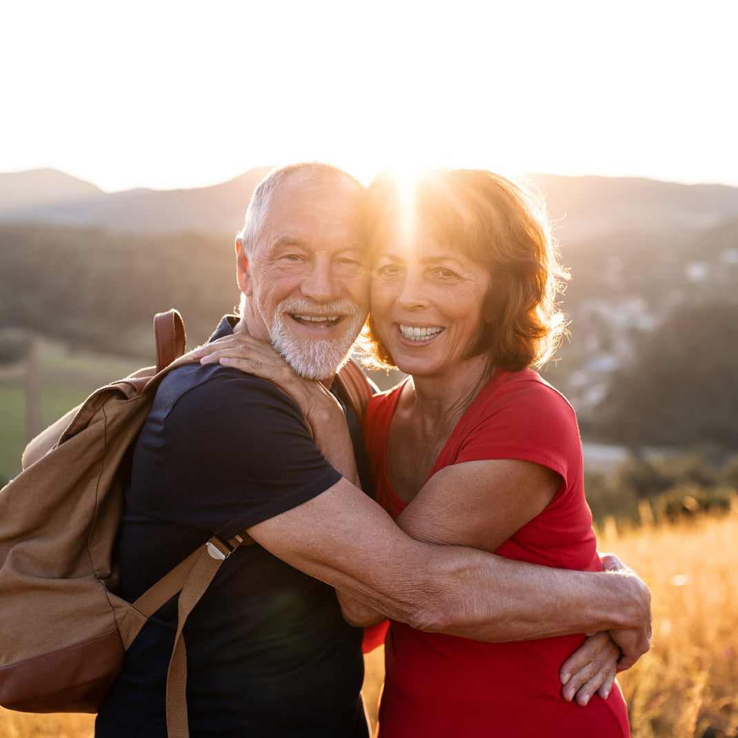 An older couple embracing outdoors at sunset, with the woman wearing a bright red top and the man wearing a dark shirt and carrying a tan backpack. Hills and warm golden light are visible in the background.