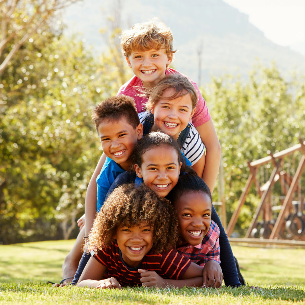 Five smiling children posing together outdoors on a grassy lawn with trees and a mountain in the background.