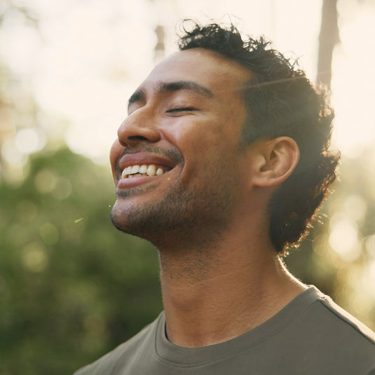 A close-up portrait of a smiling man with closed eyes, wearing a dark olive green t-shirt, set against a warm, sunlit, and blurred outdoor background.