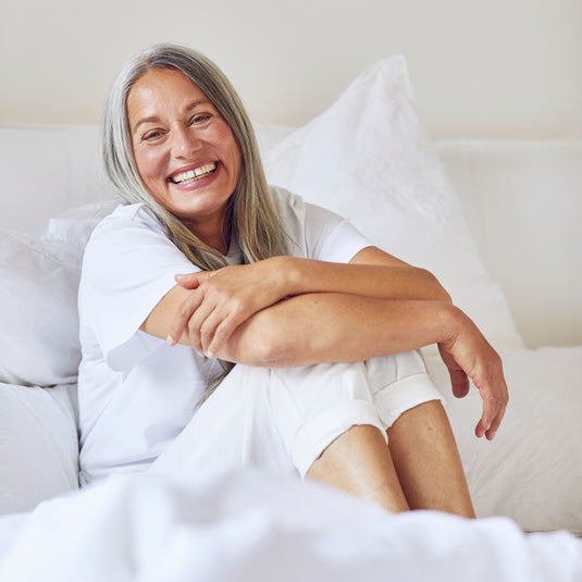 A smiling older woman with gray hair sits on a white bed with pillows, wearing a white short-sleeve top and white shorts.