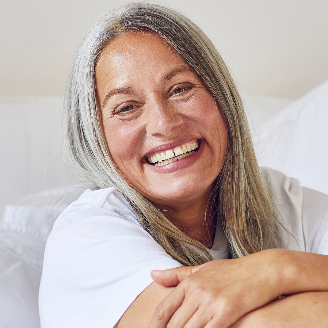 A smiling older woman with long gray hair resting her head on her arms, wearing a white t-shirt against a soft white background.