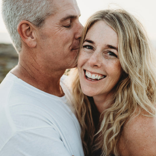 A close-up lifestyle photo of a smiling blonde woman and a kissing man in a white t-shirt, set against a warm, softly blurred outdoor background.