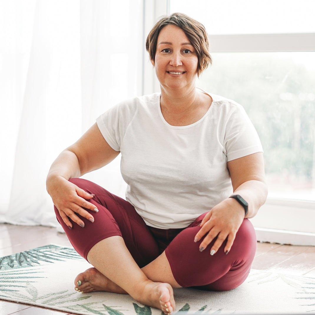 A woman with short brown hair sits cross-legged on a patterned yoga mat in a bright room. She is wearing a white short-sleeve t-shirt and burgundy leggings.