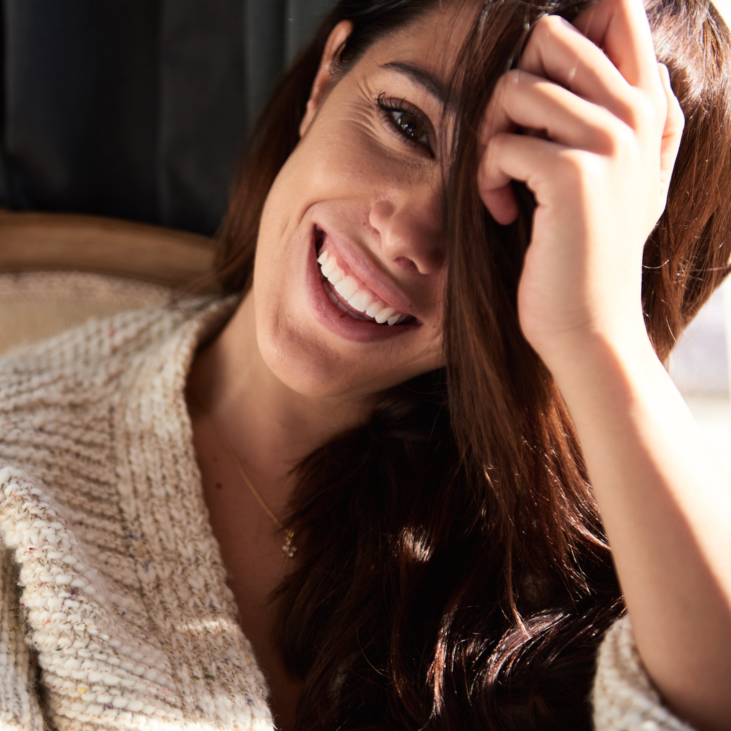 A close-up lifestyle shot of a smiling woman with long brown hair, wearing a cream-colored textured knit top, with warm sunlight illuminating her face and hair.