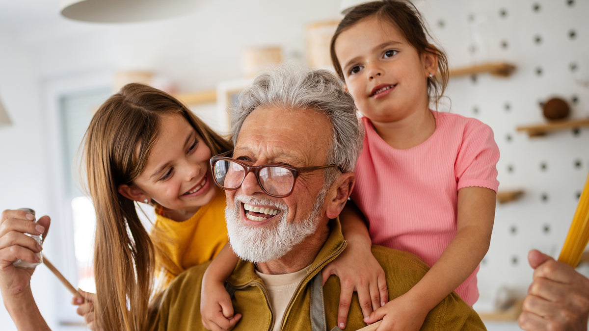 A smiling older man with gray hair and glasses carries a young girl on his shoulders while another girl in a pink shirt climbs onto his head, in a bright indoor room with shelves in the background.