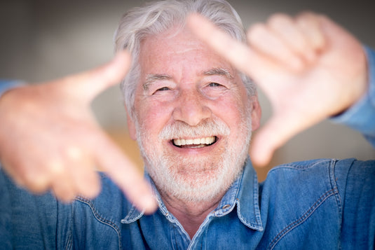 A close-up portrait of a smiling older man with gray hair and a beard, wearing a blue denim shirt and gesturing with both hands toward the camera.