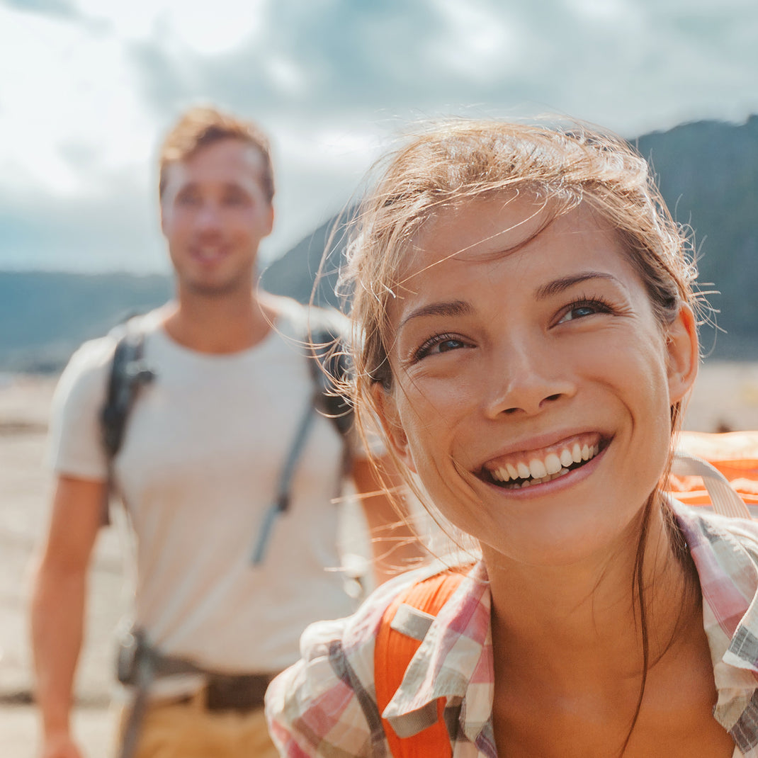 A smiling woman in the foreground wearing a plaid shirt and an orange backpack, with a blurred man in the background also wearing a backpack in an outdoor mountain setting.