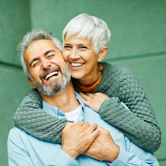An older couple embracing and laughing heartily against a solid green wall. The woman with short white hair wears a textured sage green sweater over an orange top, while the man wears a light blue button-down shirt.
