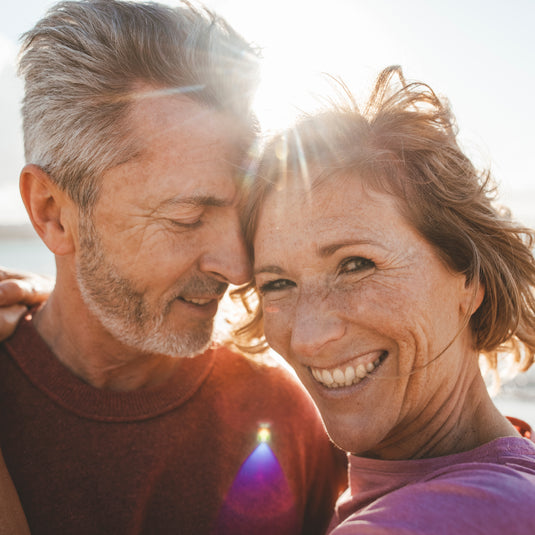 man and woman in the sunshine. The man has his eyes closed and is leaning on the woman's forehead. The woman is looking at the camera smiling.