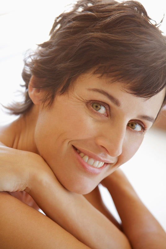 A close-up portrait of a smiling woman with short brown hair, looking directly at the camera against a bright white background.