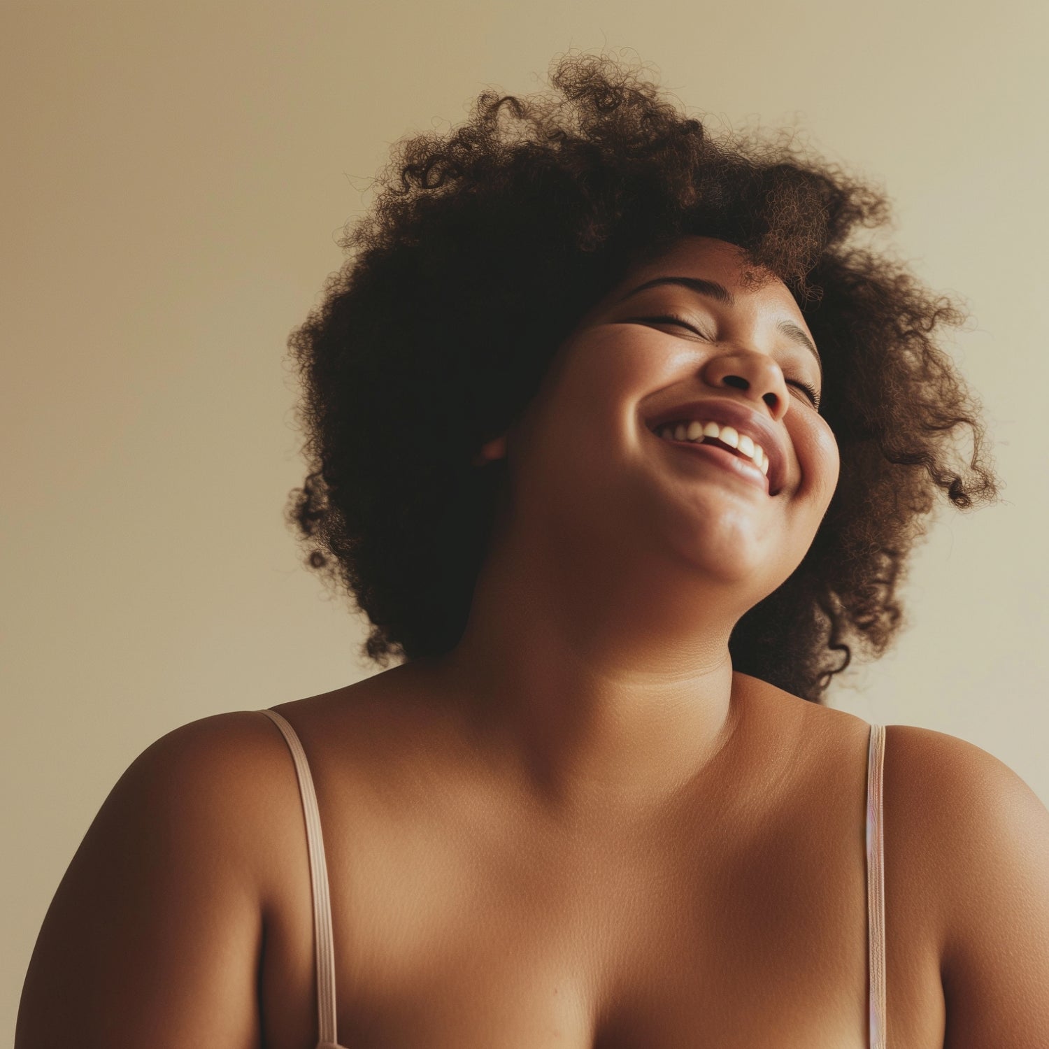 A close-up portrait of a smiling woman with voluminous dark curly hair, wearing a light beige spaghetti-strap top against a warm beige background.