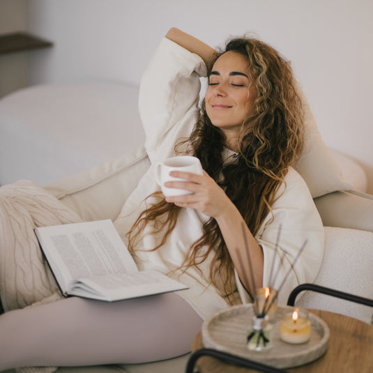 A woman with long curly brown hair relaxes on a cream sofa, holding a white mug and reading a book. She is wearing a white sweatshirt and light gray leggings, with a small wooden tray, reed diffuser, and candle on a side table in the foreground.
