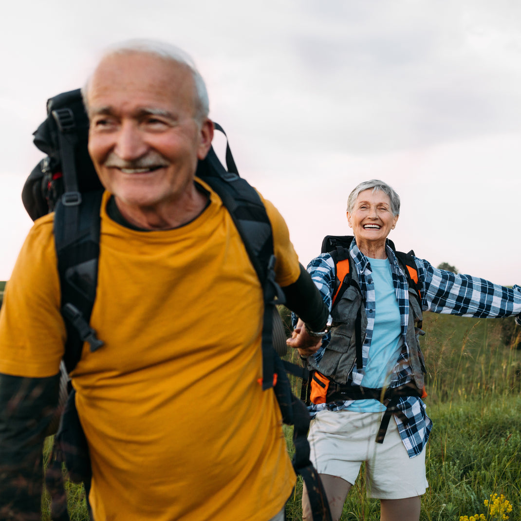 Two smiling senior hikers outdoors in a grassy field, one in the foreground wearing a yellow shirt and black backpack, and the other behind wearing a blue plaid shirt over a light blue top with a black and orange backpack.