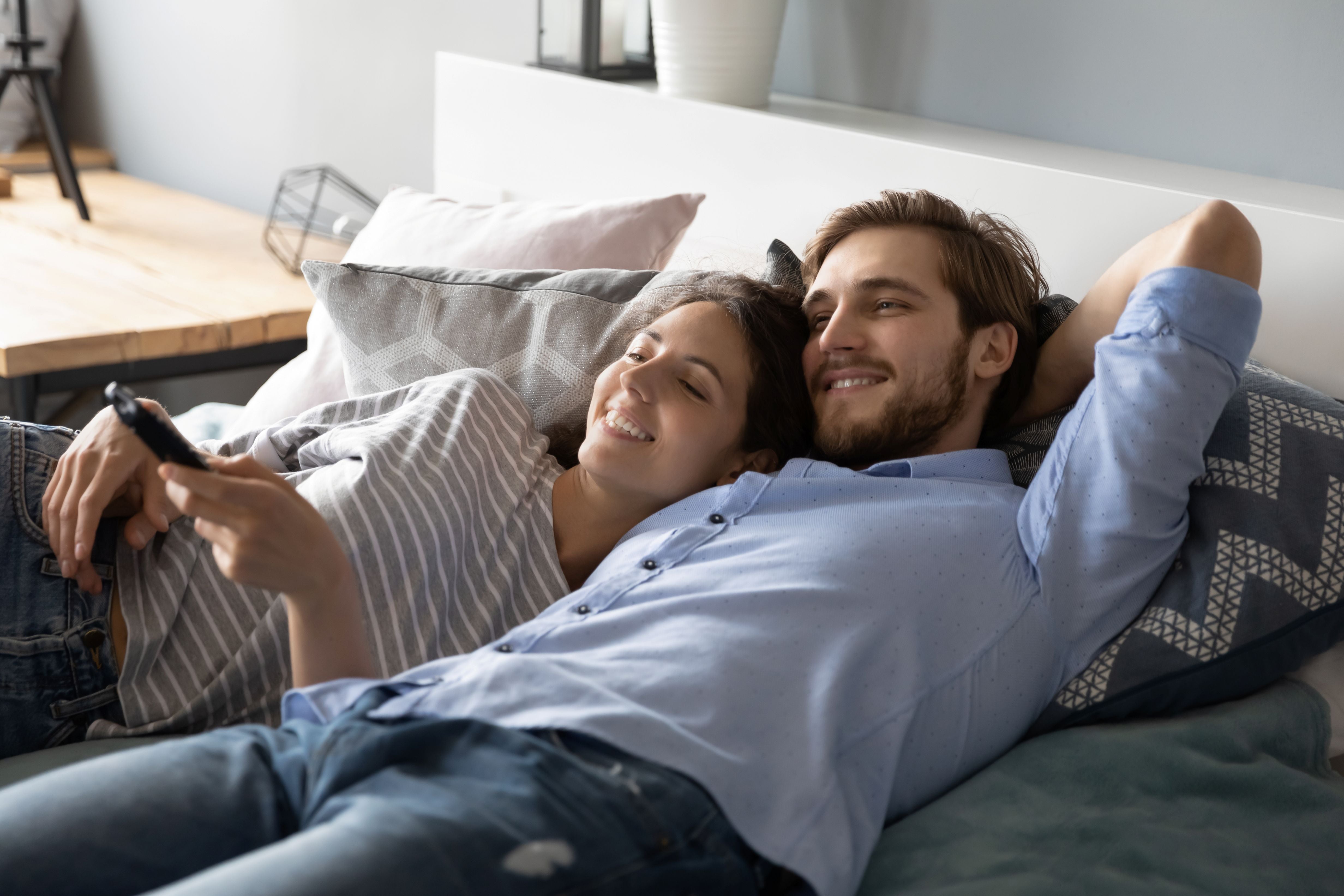 Couple lying on a couch, smiling and using a remote control.