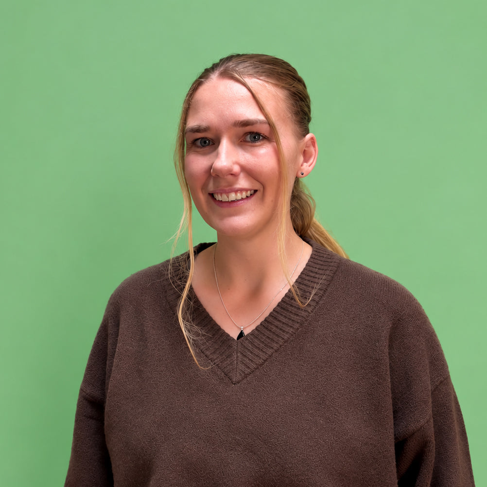 A smiling woman with blonde hair tied back, wearing an oversized brown knit V-neck sweater, standing against a solid light green background.