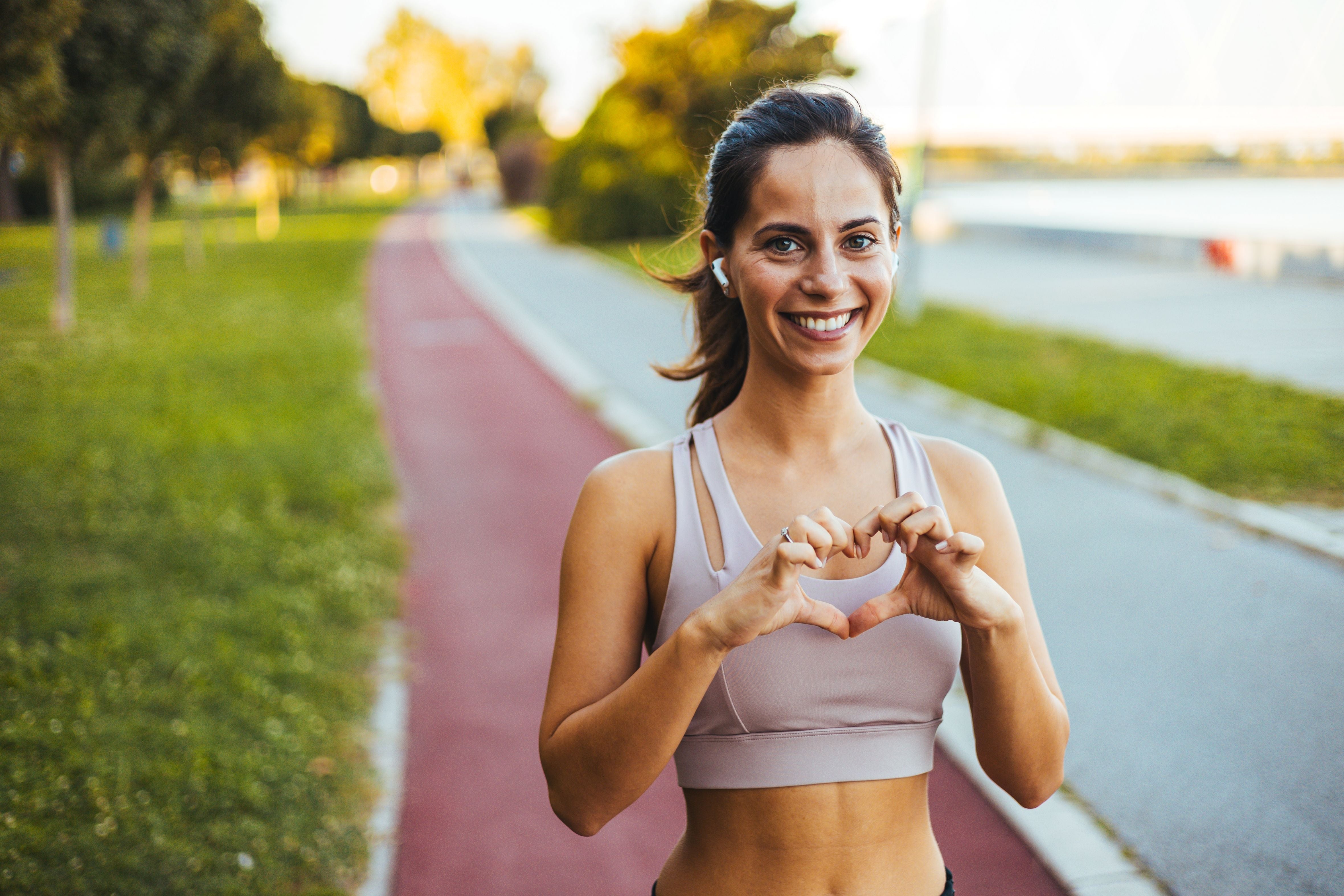 Woman in athletic wear making a heart shape with her hands on a path in a park.