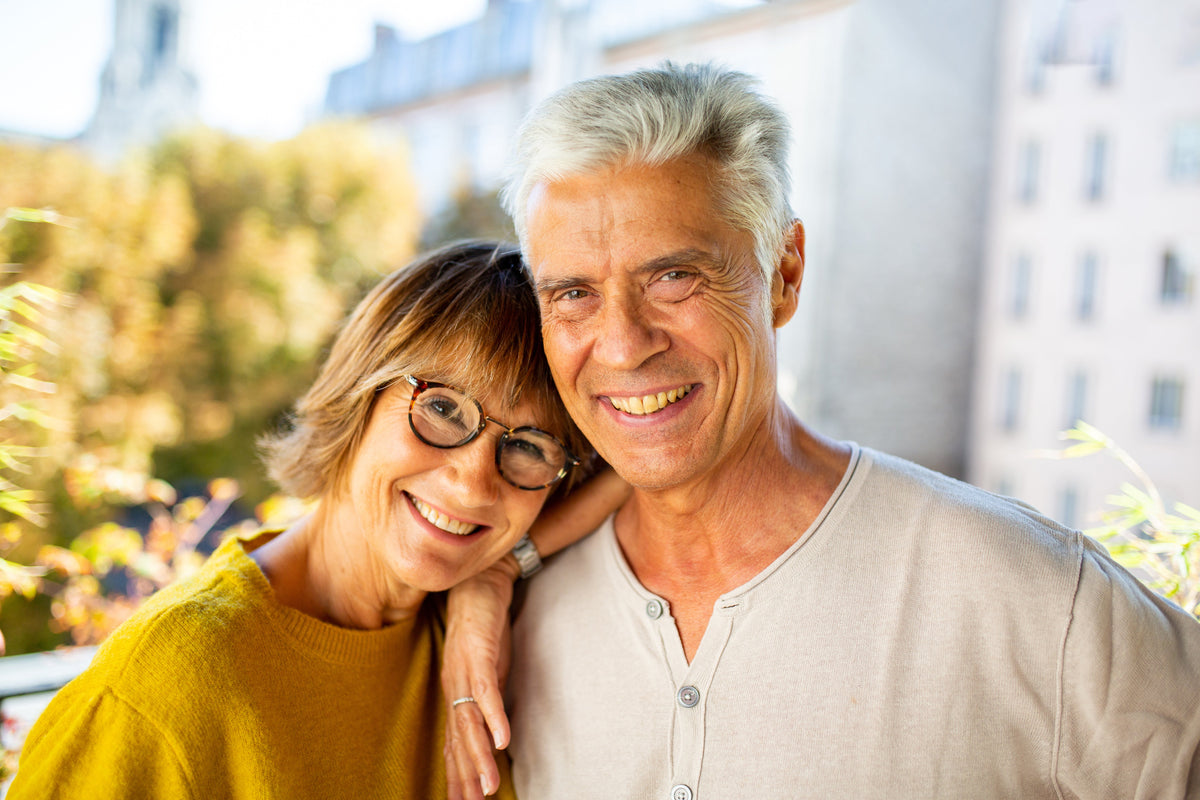An older couple smiling and posing together outdoors on a balcony, with the woman in a mustard yellow sweater and the man in a light beige henley shirt.