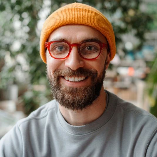 Man wearing a yellow beanie and red glasses with a blurred outdoor background
