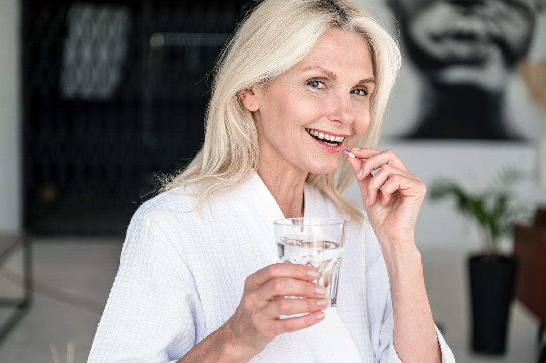 Woman in a white robe holding a glass of water and a toothbrush, with a blurred indoor background.