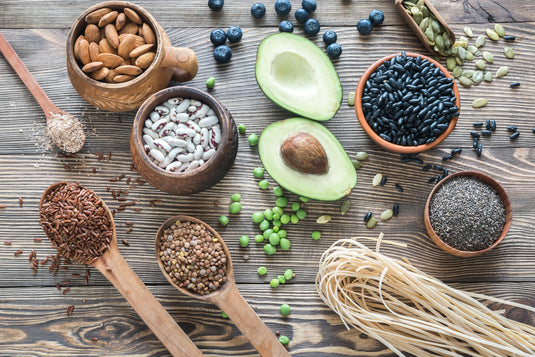 Assorted health foods including avocados, nuts, seeds, and beans on a wooden surface.
