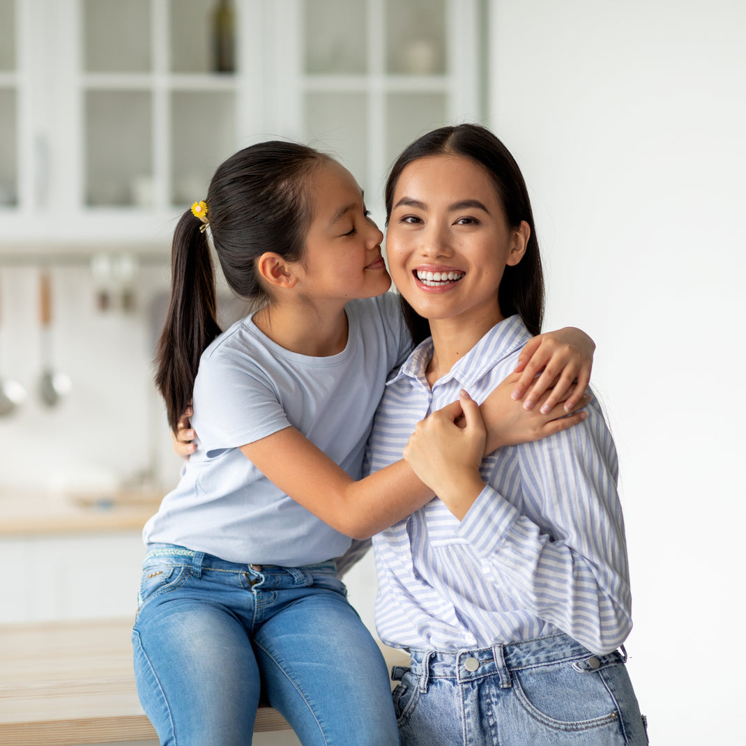A young girl with a ponytail kisses her mother on the cheek as they sit together in a bright, modern kitchen. The girl is wearing a light blue t-shirt and blue jeans, while her mother wears a light blue and white striped button-down shirt and blue jeans.