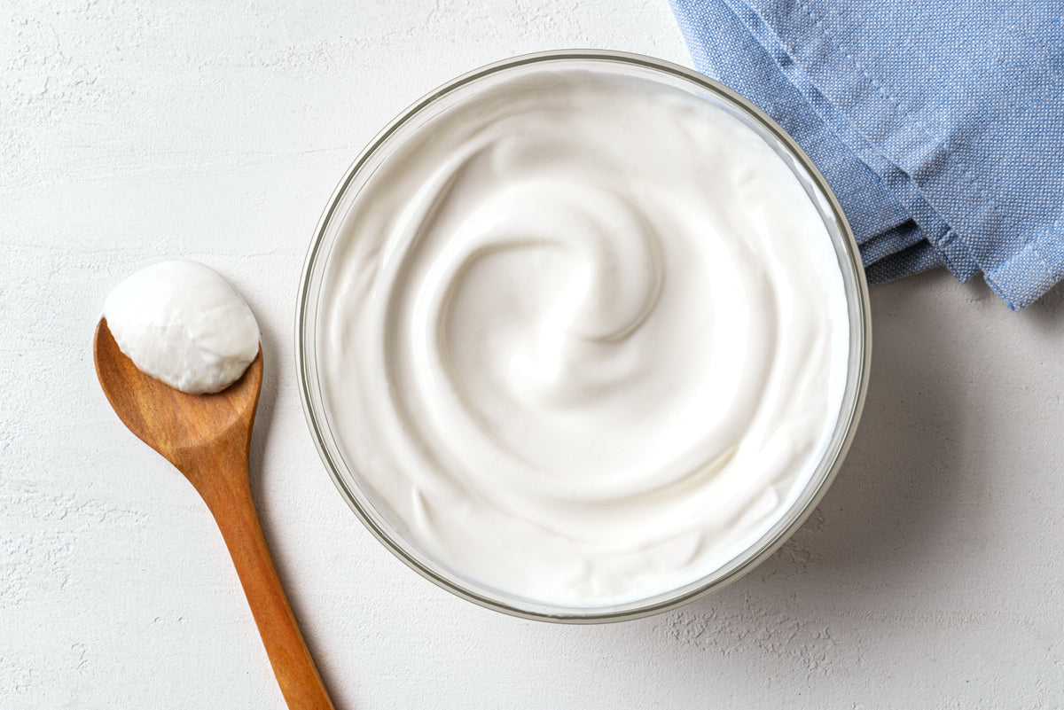 A glass bowl filled with thick white whipped cream or yogurt, shown from above on a light textured surface with a wooden spoon holding a dollop of the white substance and a folded blue cloth in the corner.